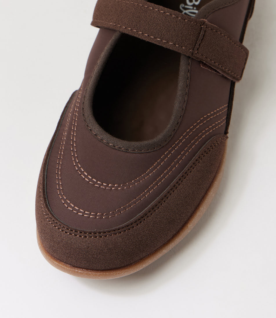 A close-up of a dark brown slip-on shoe with a hook-and-loop strap across the top, featuring decorative contrast stitching and a gum outsole, shown on a light gray background.